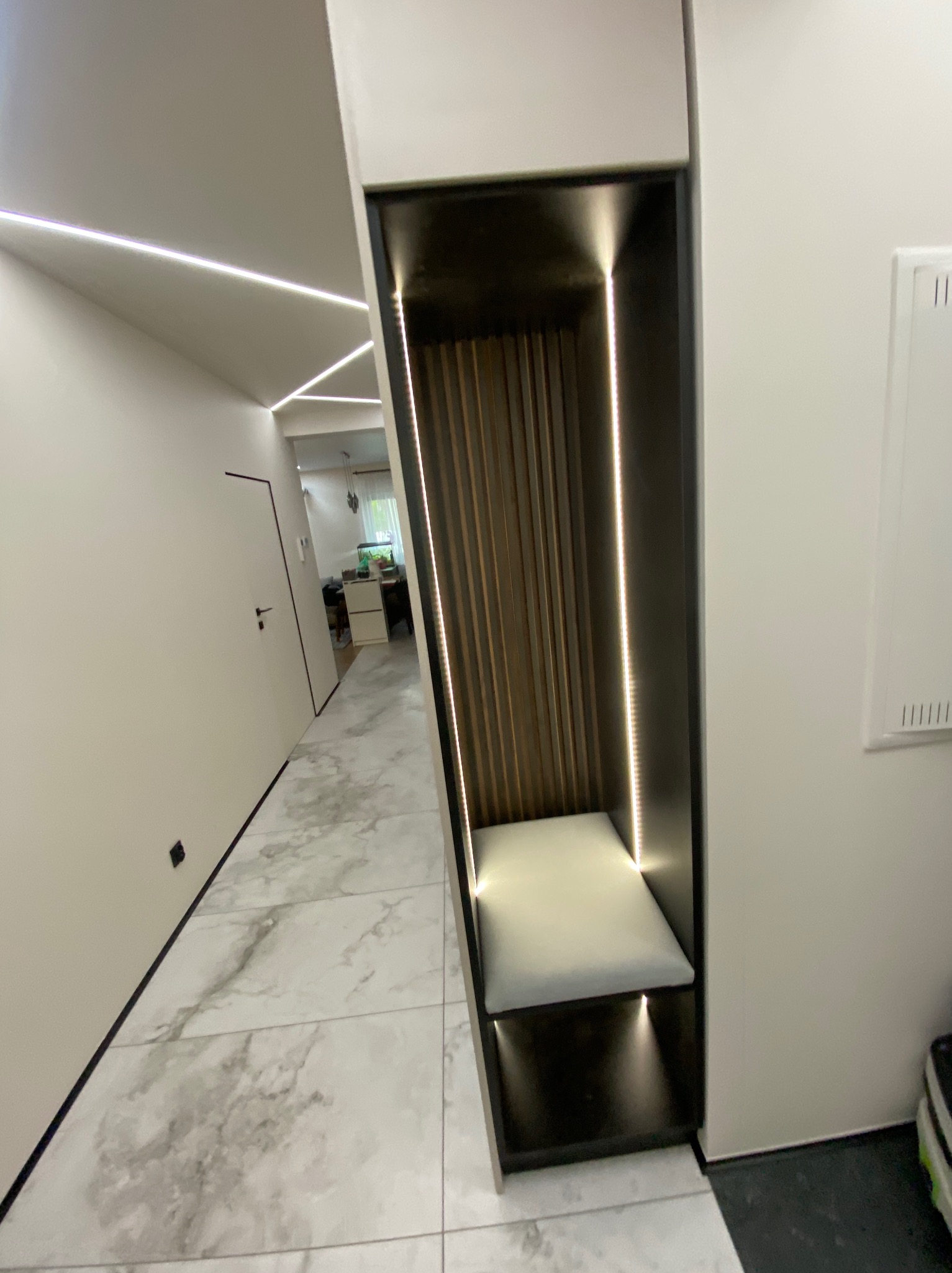 Nowoczesna zabudowa wnęki z siedziskiem i oświetleniem LED w przedpokoju. Drewniane lamele na ścianie wnęki, marmurowe płytki podłogowe, minimalistyczny design.