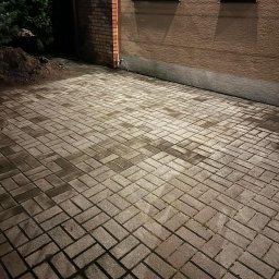 Paweł Podniesiński - Karetka Ogrodowa - Ułożona kostka brukowa w jodełkę przy budynku z oknami, z kupką ziemi obok. W rogu leży miotła z czerwonym włosiem. Nocne oświetlenie.