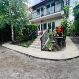 Nowy chodnik z kostki brukowej prowadzi do schodów wejściowych domu z ogrodem. Widoczne świeżo posadzone rośliny i drzewo brzozy. Ujęcie z poziomu gruntu.