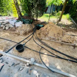 Montaż systemu nawadniania w ogrodzie: czarne rury, wykopana ziemia, częściowo ułożona kostka brukowa, narzędzia ogrodnicze i worki z materiałami.