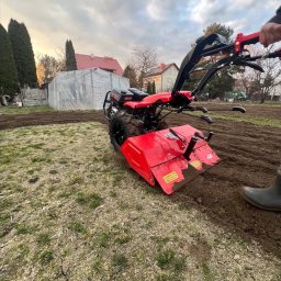 Paweł Podniesiński - Karetka Ogrodowa - Czerwony glebogryzarek w trakcie pracy na polu, przygotowując ziemię pod uprawę. W tle widoczny szklarnia i zabudowania.