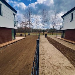 Paweł Podniesiński - Karetka Ogrodowa - Nierówny teren przygotowywany pod trawnik między dwoma nowoczesnymi domami. Widoczne ślady po maszynie do wyrównywania ziemi i pracujące osoby w oddali.