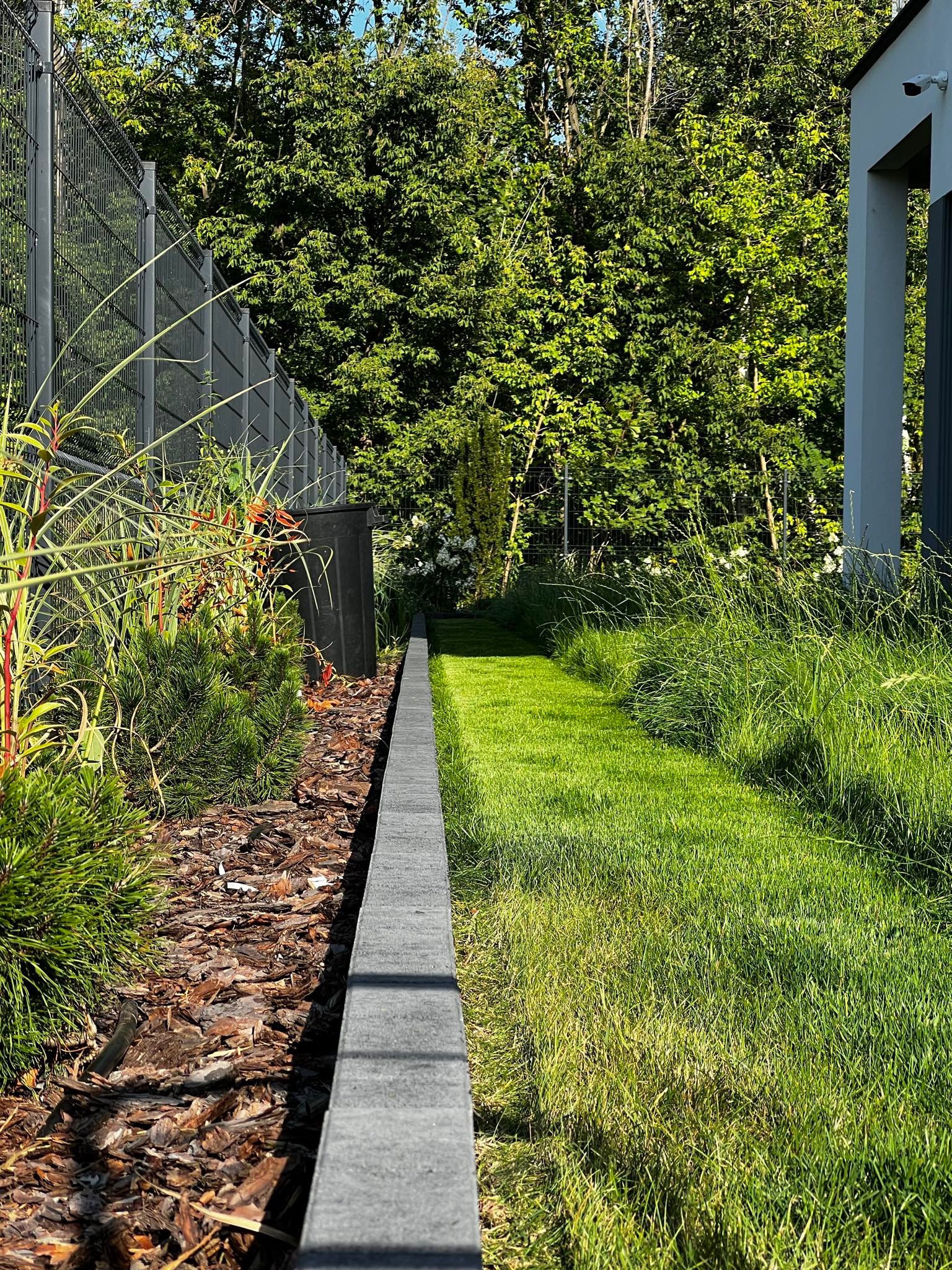 Starannie zaprojektowany ogród z wytyczoną ścieżką z kostki obrzeżowej, oddzielającą rabatę z korą i roślinami ozdobnymi od trawnika, z widocznym ogrodzeniem panelowym i fragmentem budynku w tle.
