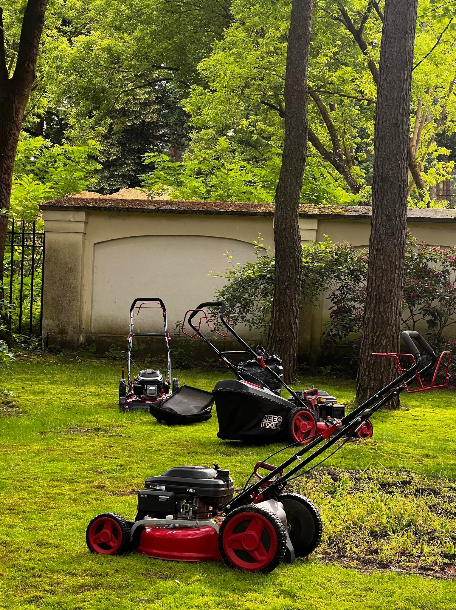 Trzy kosiarki do trawy, w tym jedna marki MEEC TOOLS, ustawione na trawniku w ogrodzie z drzewami i murem w tle.