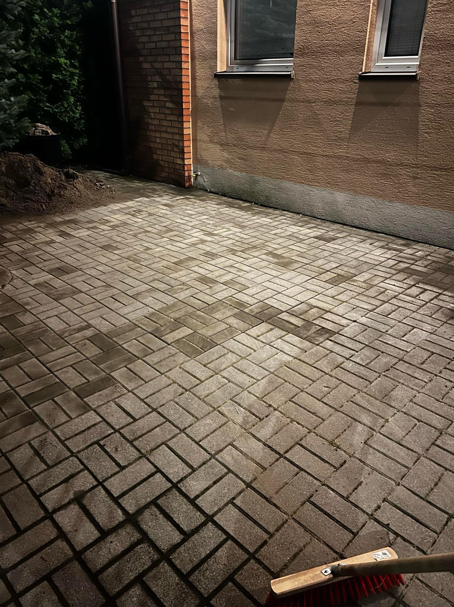 Ułożona kostka brukowa w jodełkę przy budynku z oknami, z kupką ziemi obok. W rogu leży miotła z czerwonym włosiem. Nocne oświetlenie.