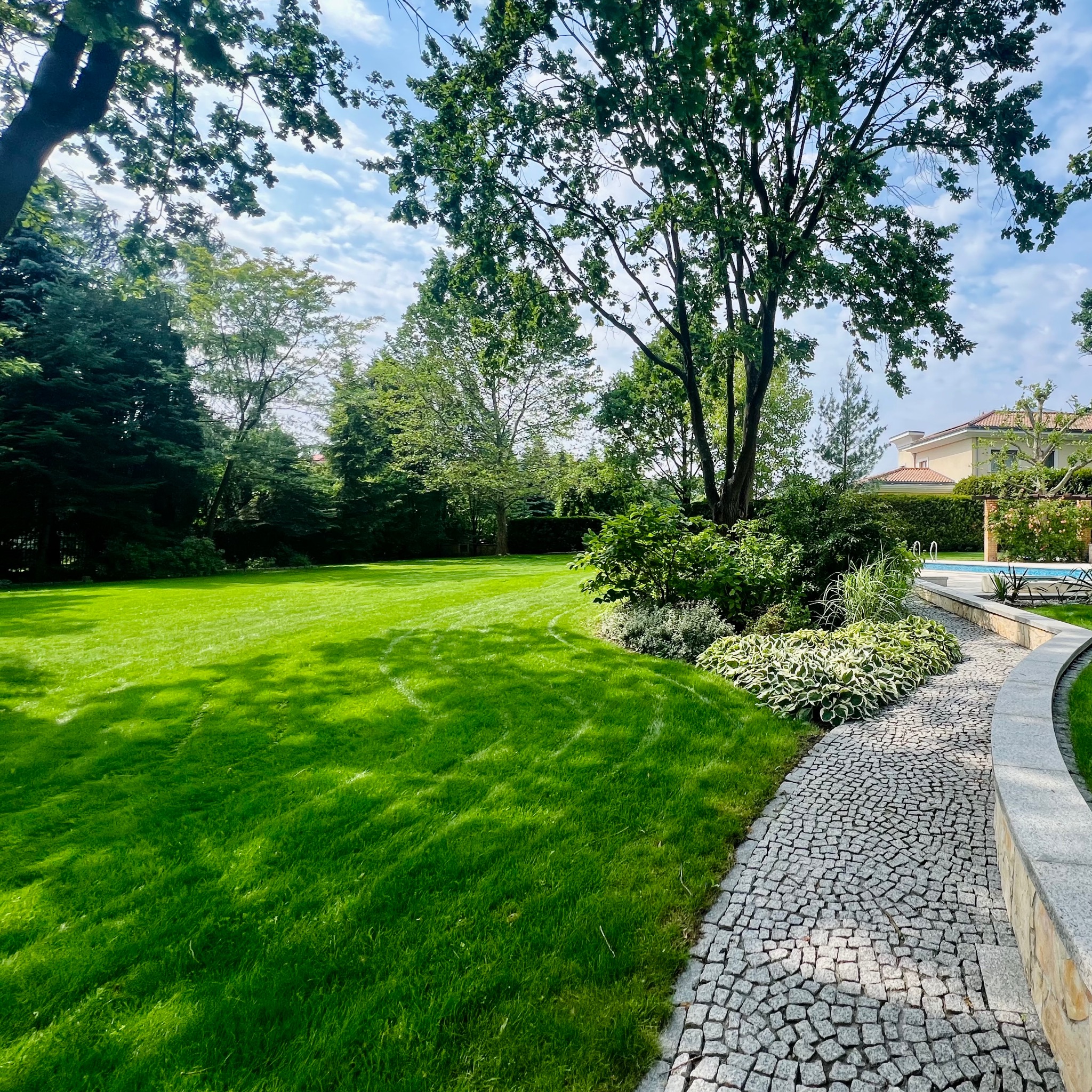 Zadbany ogród z równo skoszoną trawą, ozdobnymi rabatami i kamienną ścieżką. W tle widoczny budynek z basenem, całość w słoneczny dzień.