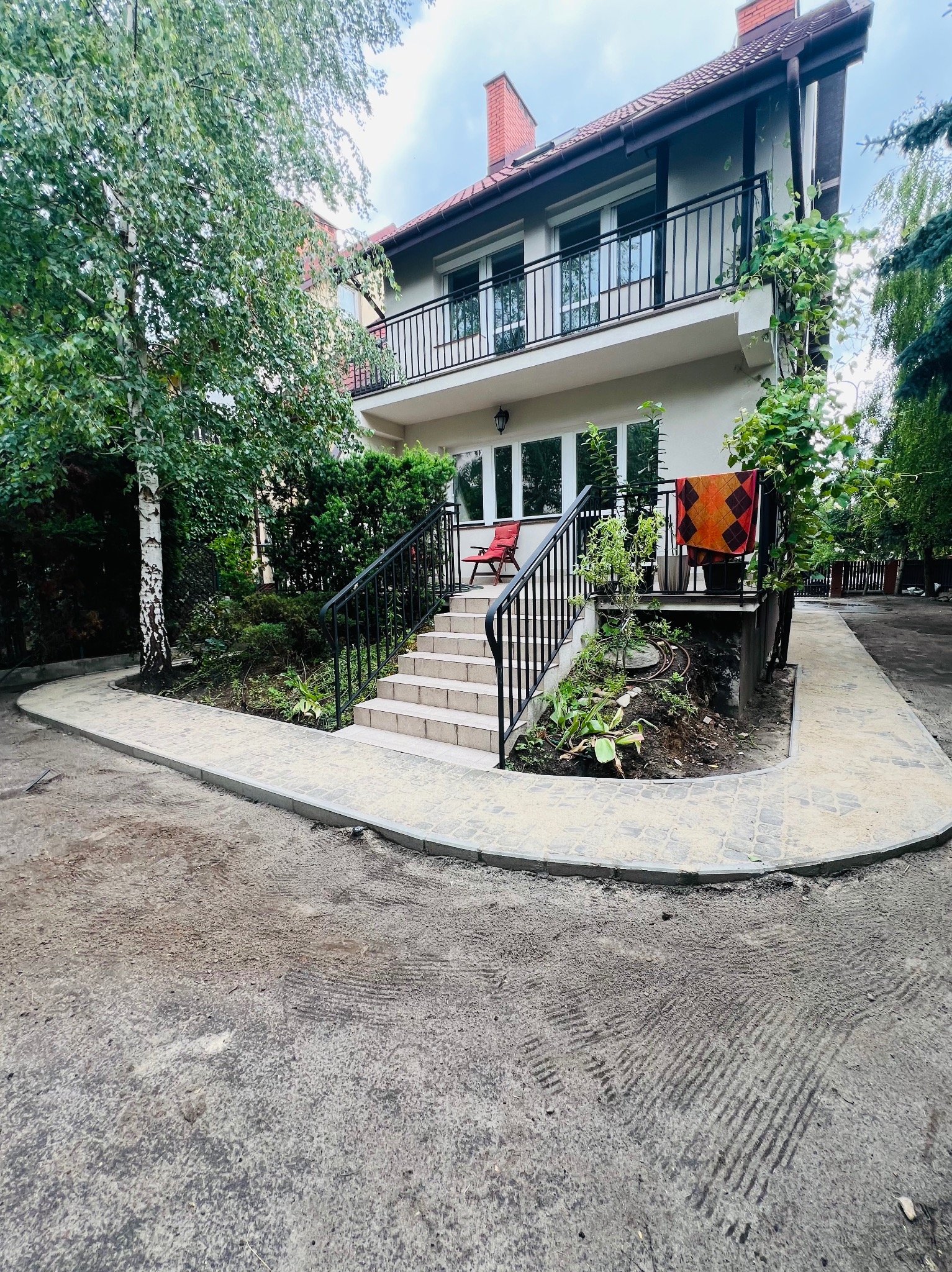 Nowy chodnik z kostki brukowej prowadzi do schodów wejściowych domu z ogrodem. Widoczne świeżo posadzone rośliny i drzewo brzozy. Ujęcie z poziomu gruntu.