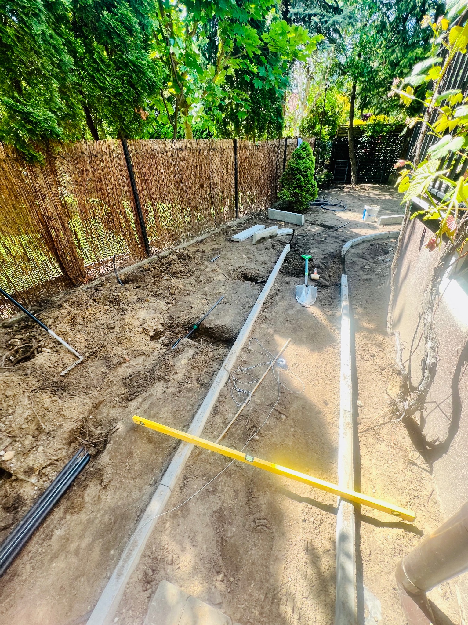 Przygotowanie terenu pod ścieżkę w ogrodzie: wykopana ziemia, obrzeża betonowe, poziomica, narzędzia ogrodnicze i rury, tło z roślinnością i płotem z trzciny.