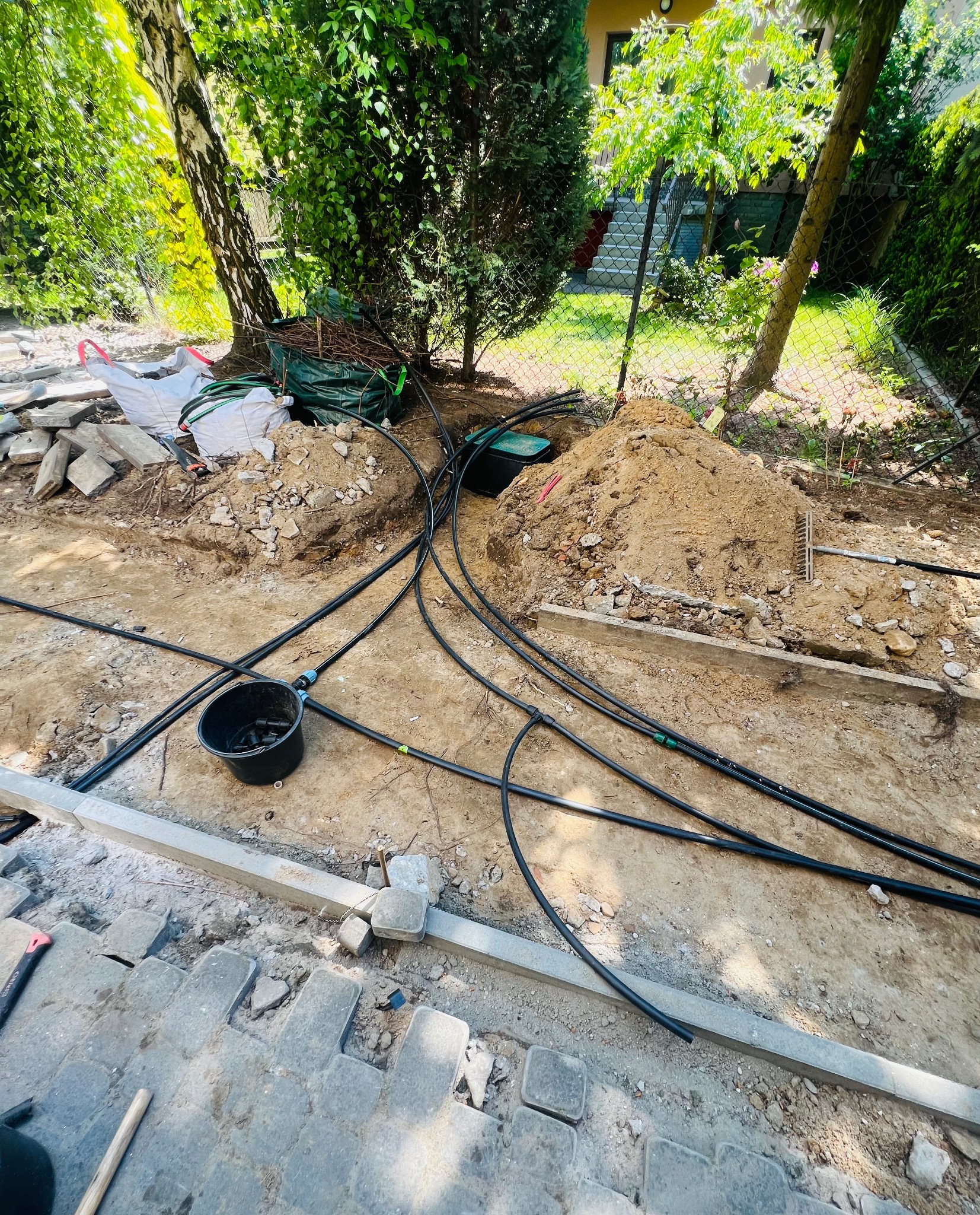 Montaż systemu nawadniania w ogrodzie: czarne rury, wykopana ziemia, częściowo ułożona kostka brukowa, narzędzia ogrodnicze i worki z materiałami.