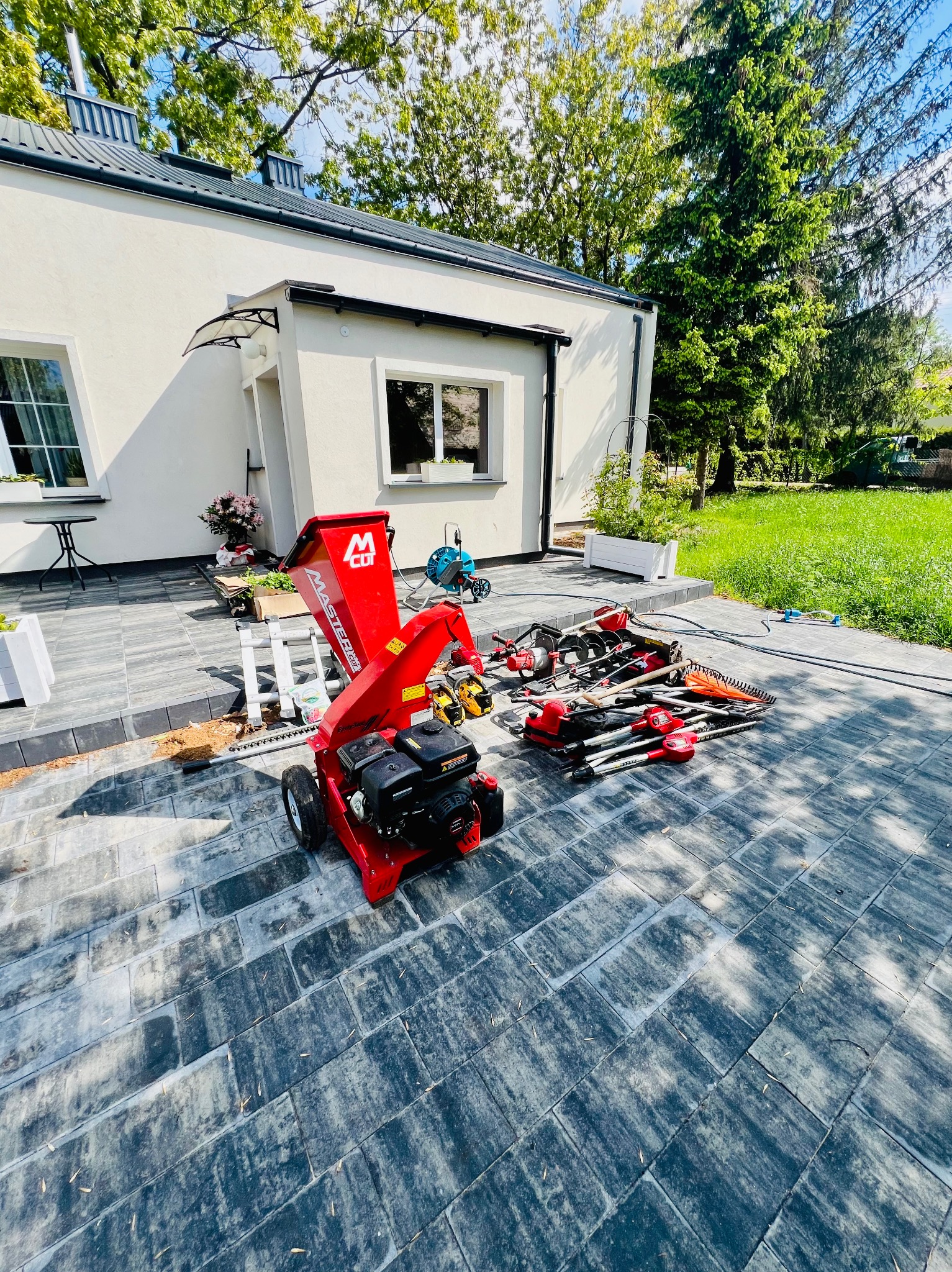 Czerwony rozdrabniacz gałęzi i narzędzia ogrodowe na szarym brukowanym podjeździe obok białego domu z zielonym trawnikiem w tle.