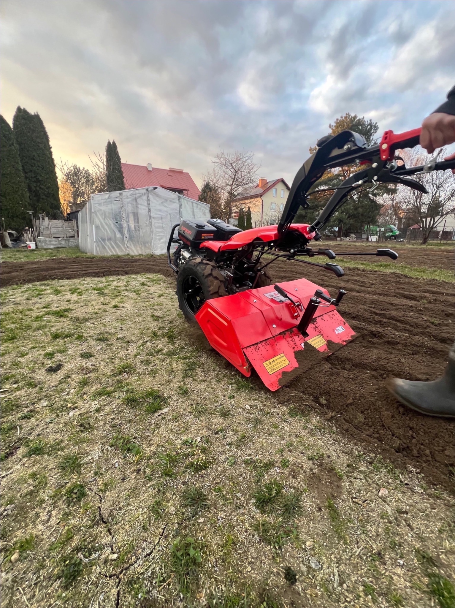 Czerwony glebogryzarek w trakcie pracy na polu, przygotowując ziemię pod uprawę. W tle widoczny szklarnia i zabudowania.