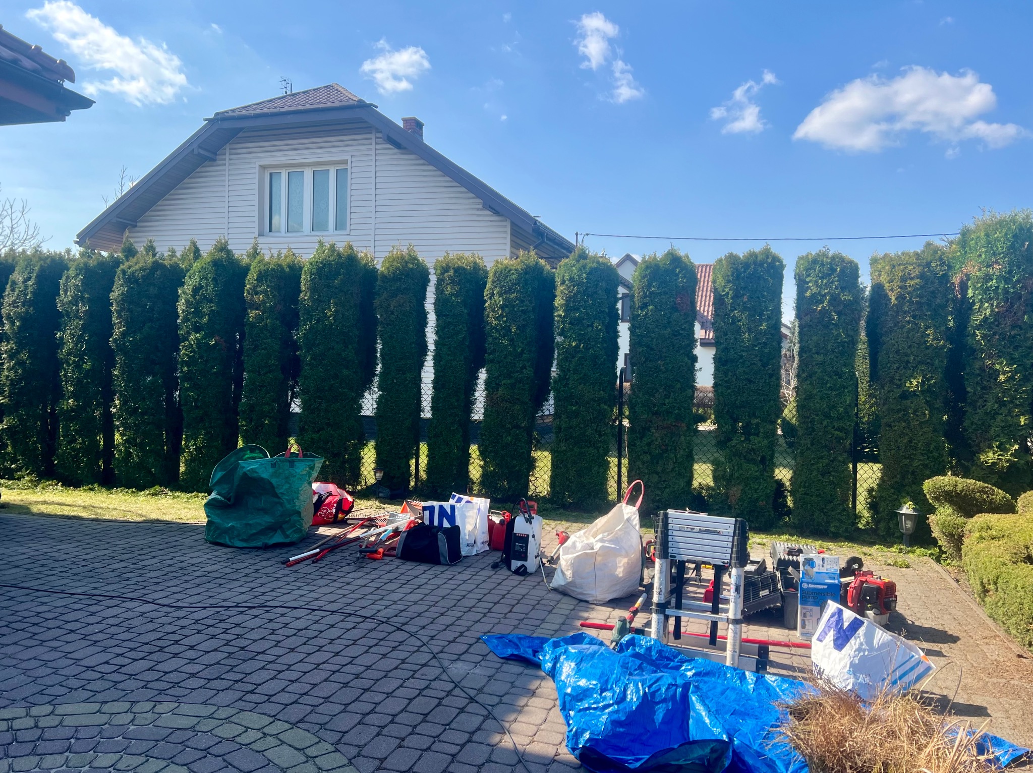 Sprzęt ogrodniczy rozłożony na brukowanym podwórku w Pruszkowie, gotowy do pracy. W tle rząd tuj i dom jednorodzinny w słoneczny dzień.