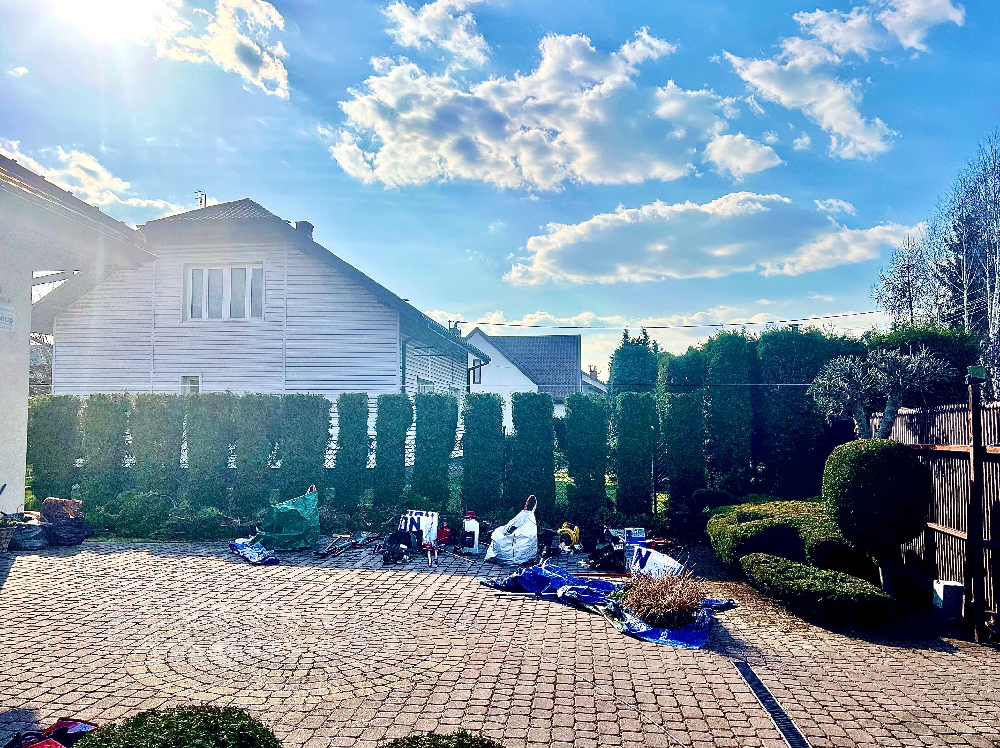 Porządkowanie po przycinaniu żywopłotu z tuj. Widoczne narzędzia, worki na odpady i resztki gałęzi na brukowanym podjeździe przed domem w słoneczny dzień.