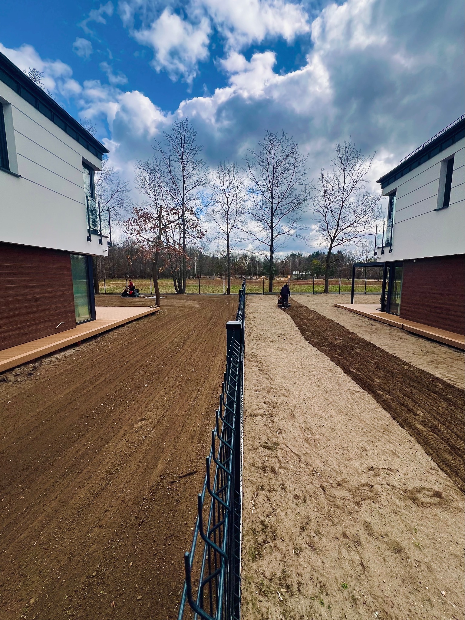 Nierówny teren przygotowywany pod trawnik między dwoma nowoczesnymi domami. Widoczne ślady po maszynie do wyrównywania ziemi i pracujące osoby w oddali.