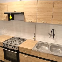 Yurii Cherednychenko - Wykończona kuchnia z jasnymi drewnianymi szafkami, białymi kafelkami na ścianie, metalowym zlewem i kuchenką gazową, widoczny grzejnik pod oknem i zawieszka zapachowa pod okapem.