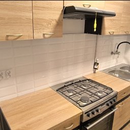 Yurii Cherednychenko - Jasna kuchnia po remoncie w Kwidzynie: białe, prostokątne płytki na ścianie, drewnopodobne szafki górne i dolne z uchwytami, dwukomorowy zlew, kuchenka gazowa z piekarnikiem i czarny okap.