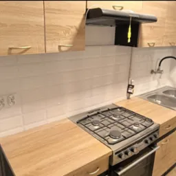 Jasna kuchnia po remoncie w Kwidzynie: białe, prostokątne płytki na ścianie, drewnopodobne szafki górne i dolne z uchwytami, dwukomorowy zlew, kuchenka gazowa z piekarnikiem i czarny okap.