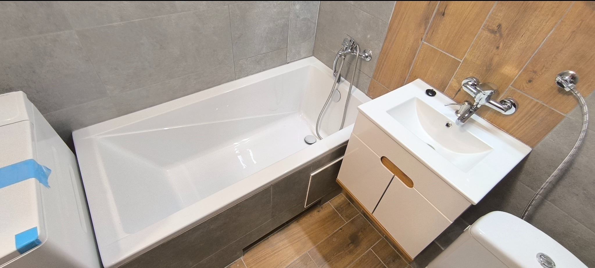 Nowoczesna łazienka po remoncie: biała wanna obudowana szarymi płytkami, umywalka z szafką w kolorze złamanej bieli z drewnianymi akcentami, drewnopodobne płytki na ścianie, widoczna toaleta...