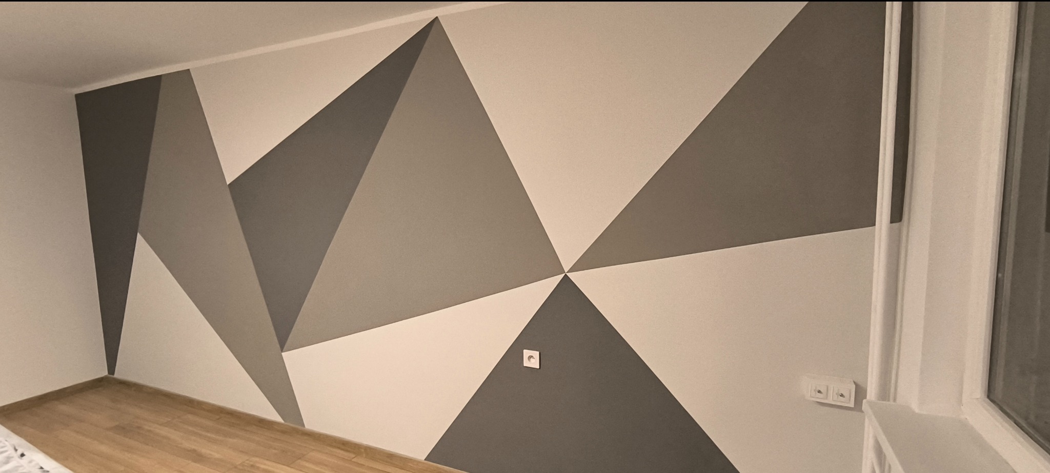 Ściana w pokoju pomalowana w geometryczne wzory z użyciem szarości i bieli, widoczne gniazdka elektryczne i fragment drewnianej podłogi.