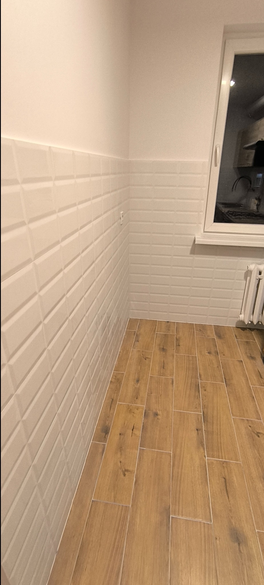 Wykończony narożnik łazienki z białymi, trójwymiarowymi płytkami ściennymi ułożonymi do połowy wysokości i drewnopodobną podłogą z płytek, widoczne gniazdko elektryczne i fragment okna z grzejnikiem.