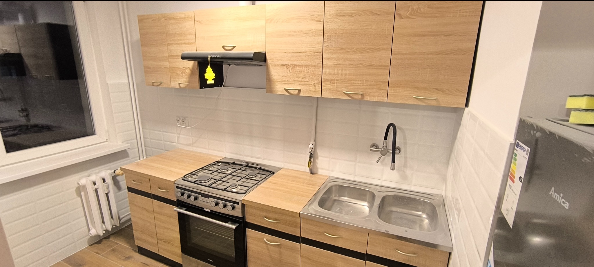 Wykończona kuchnia z jasnymi drewnianymi szafkami, białymi kafelkami na ścianie, metalowym zlewem i kuchenką gazową, widoczny grzejnik pod oknem i zawieszka zapachowa pod okapem.