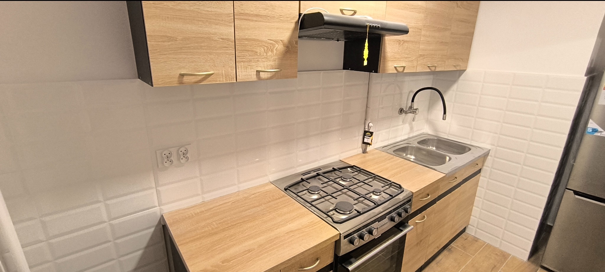 Jasna kuchnia po remoncie w Kwidzynie: białe, prostokątne płytki na ścianie, drewnopodobne szafki górne i dolne z uchwytami, dwukomorowy zlew, kuchenka gazowa z piekarnikiem i czarny okap.