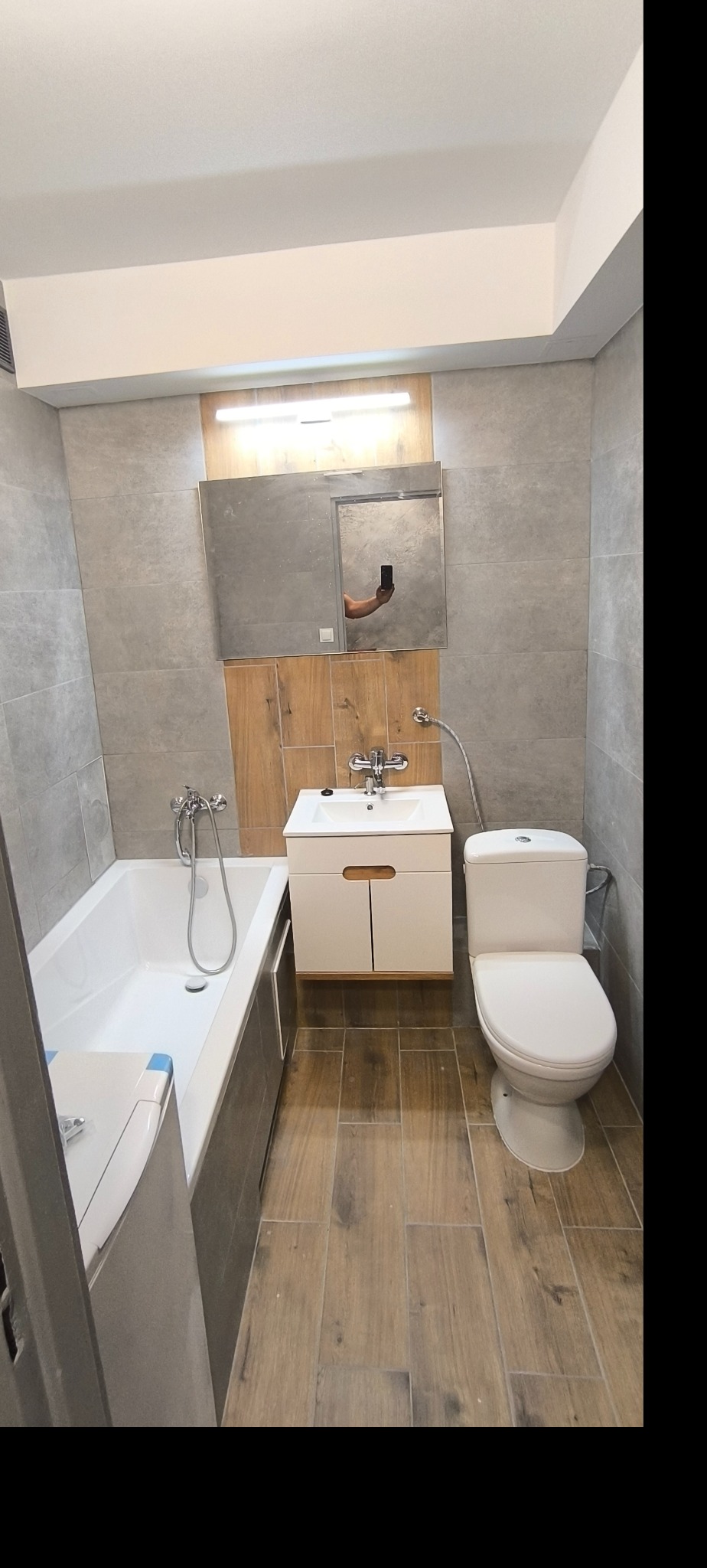 Wykończona łazienka z wanną, umywalką z szafką i toaletą, szare płytki ścienne i podłogowe imitujące drewno, lustro z oświetleniem LED, odbicie ręki fotografa w lustrze.