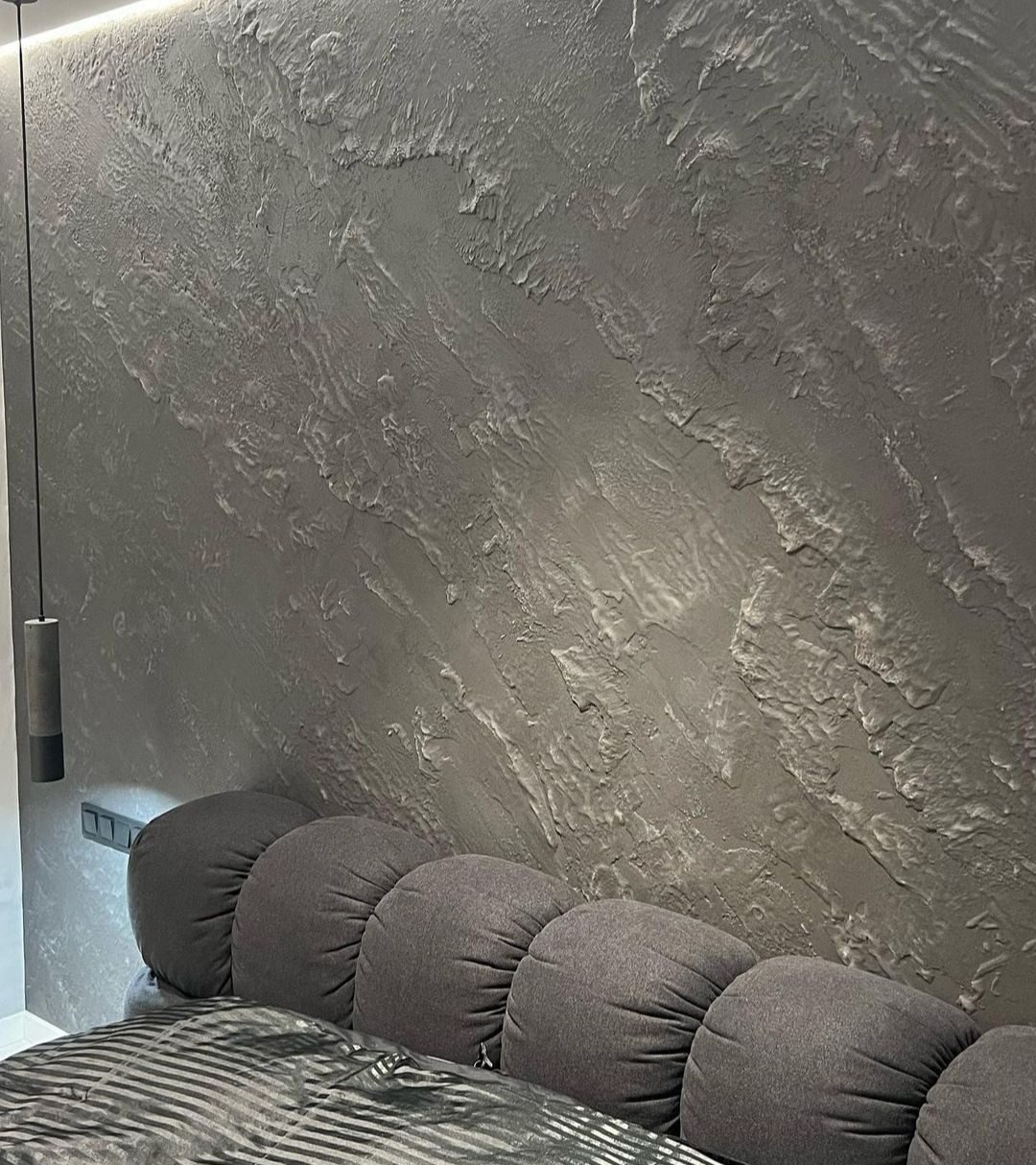 Dekoracyjna, szara ściana z efektem strukturalnym nad wezgłowiem łóżka z okrągłymi poduszkami, oświetlona listwą LED.