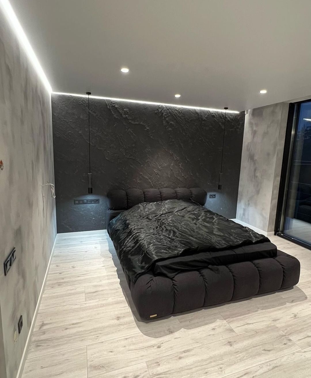 Sypialnia z łóżkiem na niskiej platformie, ściana pokryta ciemną, strukturalną gładzią, oświetlenie LED podsufitowe, panele podłogowe w odcieniach szarości.