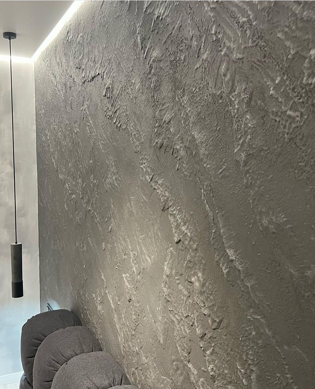 Szara ściana z efektem dekoracyjnym przypominającym naturalny kamień, oświetlona listwą LED pod sufitem i lampą wiszącą na czarnym kablu, z widocznymi okrągłymi poduszkami w dolnej części kadru.