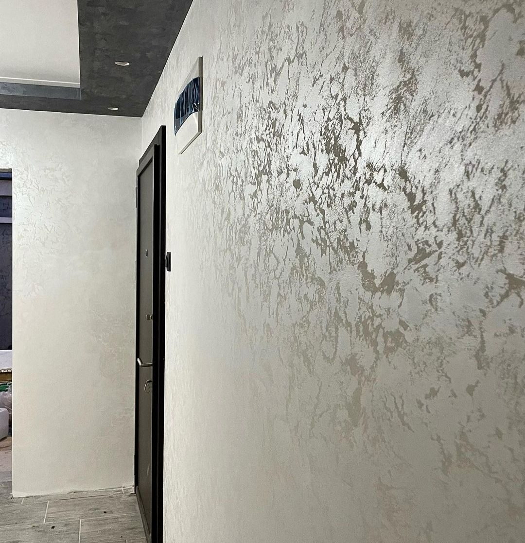 Ściana wykończona dekoracyjną, srebrzystą gładzią w korytarzu, widok na drzwi wejściowe i fragment sufitu z oświetleniem punktowym.