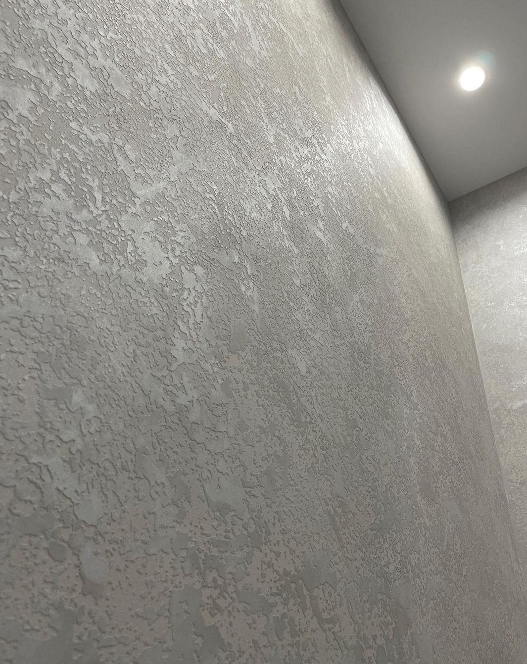 Ściana pokryta szarą, dekoracyjną strukturą z widocznym oświetleniem sufitowym punktowym.