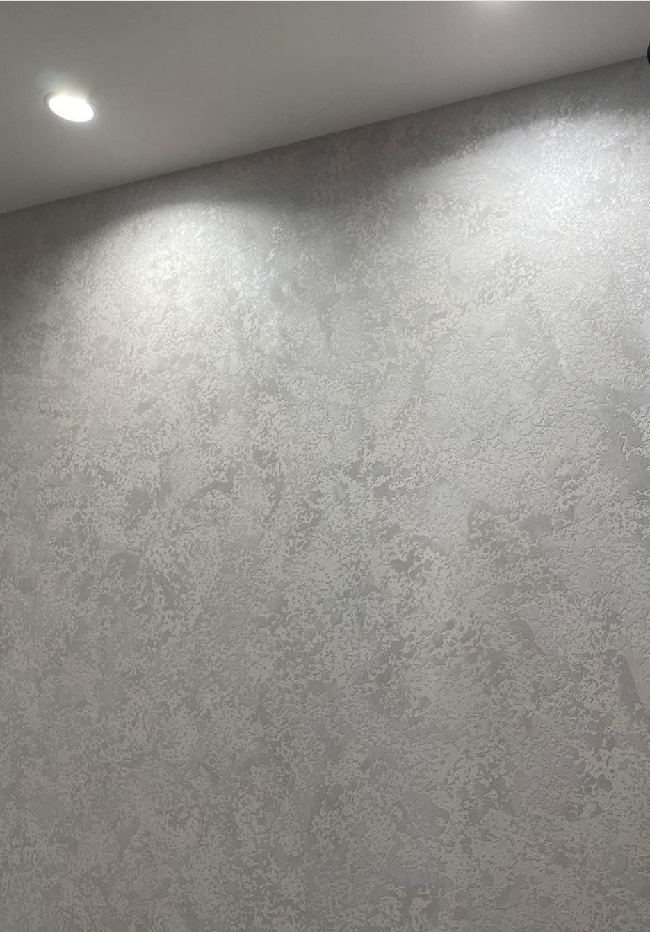 Ściana pokryta dekoracyjną, strukturalną szarą farbą, oświetlona punktowym światłem z sufitu.