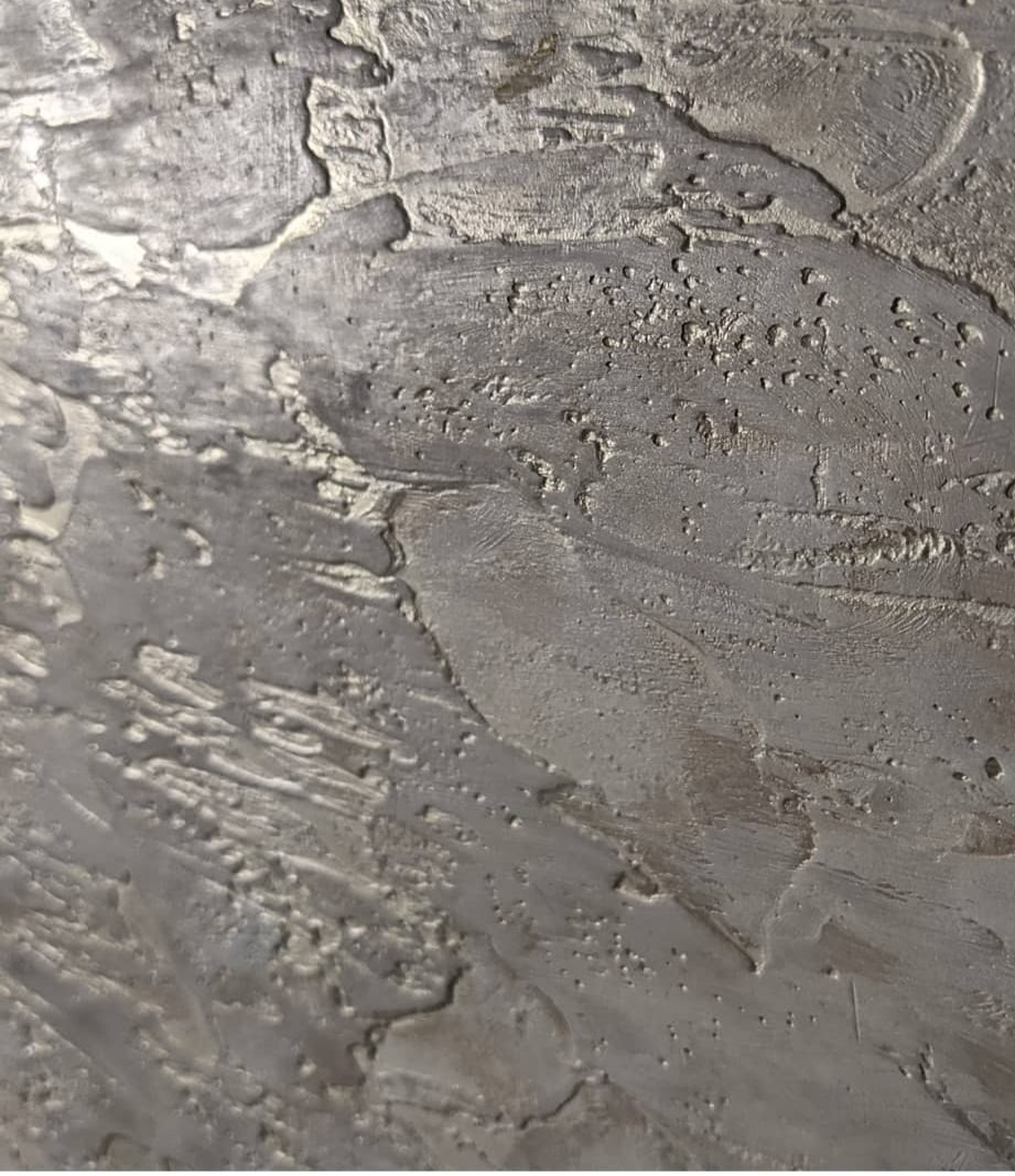 Szara, strukturalna gładź dekoracyjna na ścianie, z wyraźnymi śladami pędzla i drobnymi nierównościami, tworząca efekt surowego, industrialnego wykończenia.