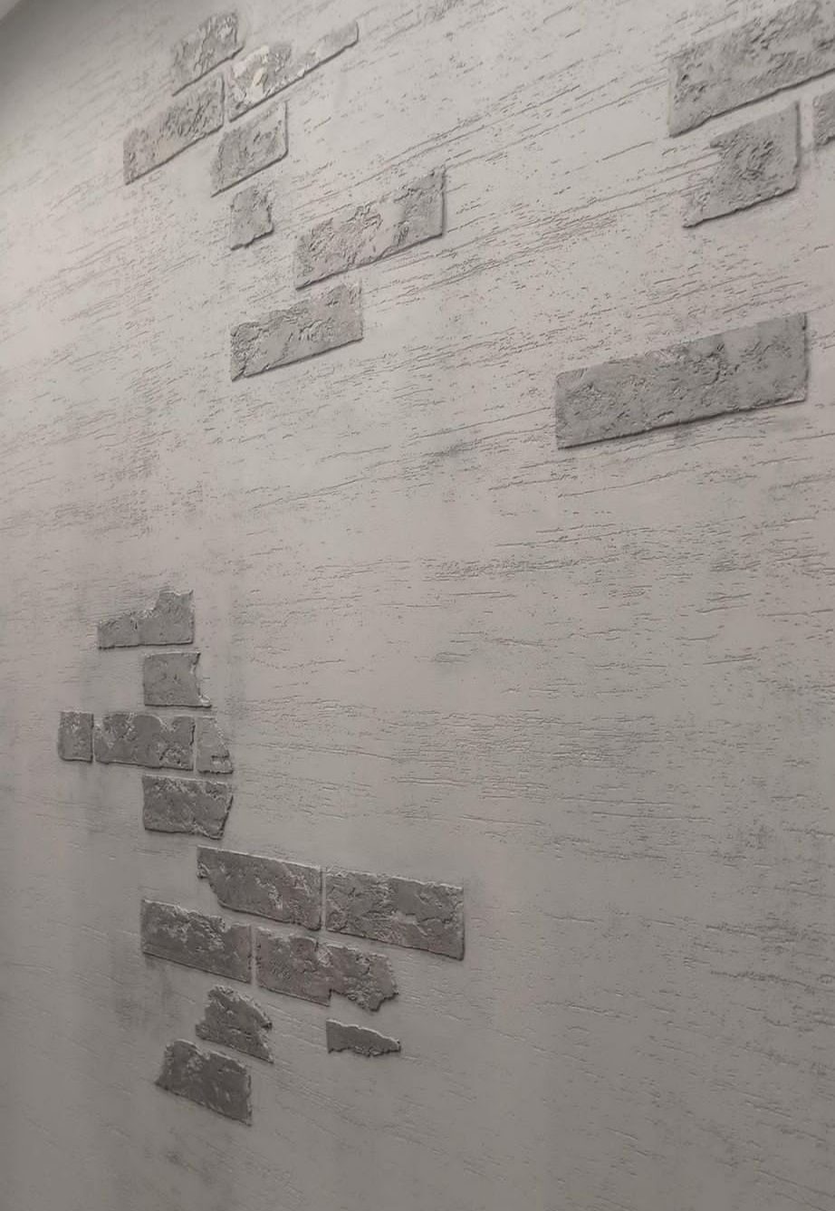 Szara ściana z dekoracyjnymi elementami imitującymi cegły, ułożonymi w nieregularny wzór na teksturowanej powierzchni.