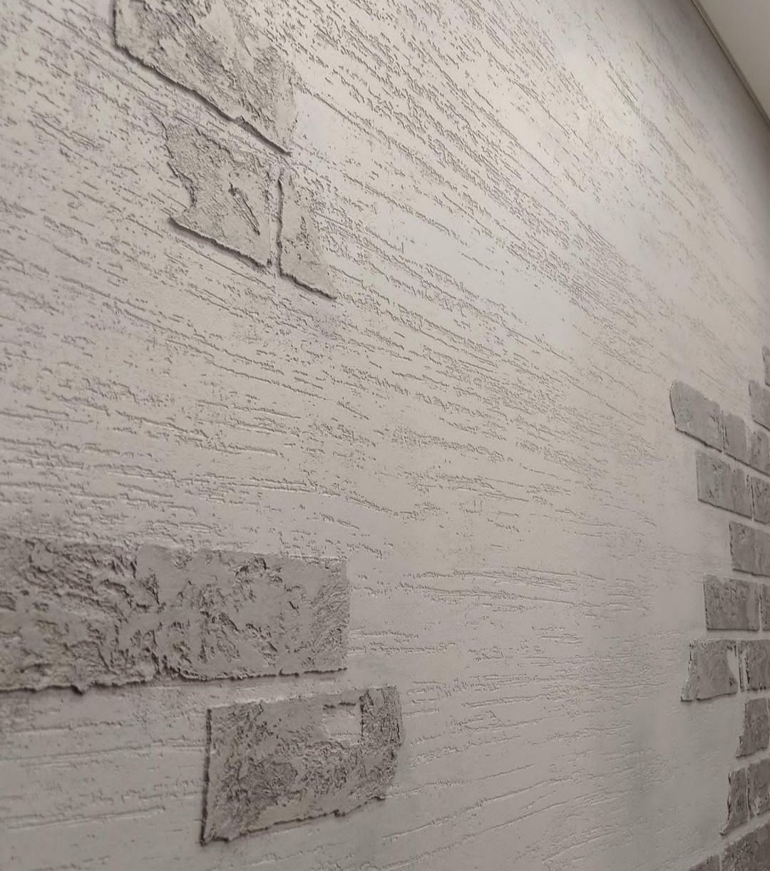 Szara ściana z dekoracyjną fakturą przypominającą cegły i poziomymi żłobieniami, imitująca surowy mur.