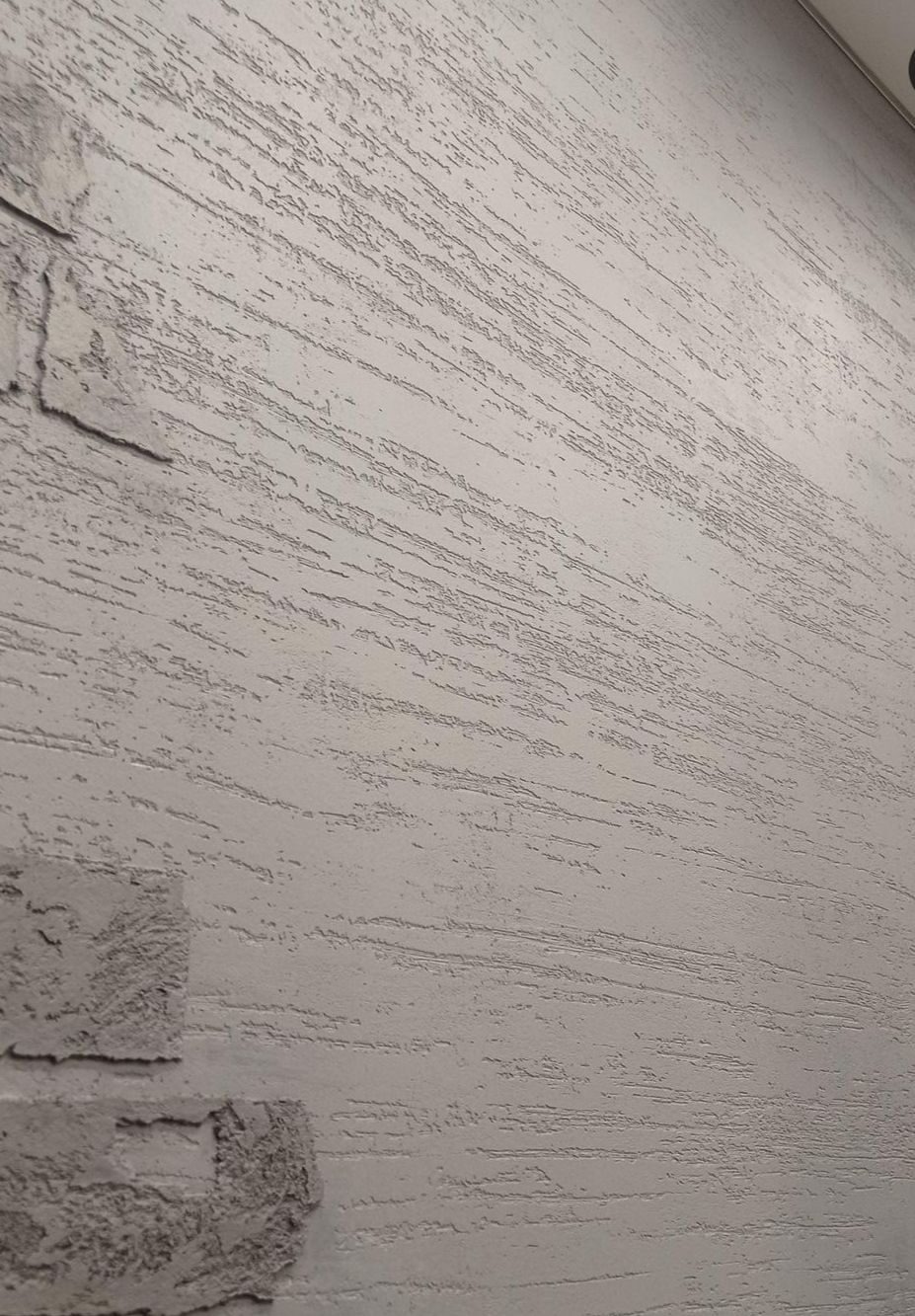 Szara ściana z efektem dekoracyjnym imitującym drewno, z fragmentem dekoracji przypominającej surową cegłę w lewym dolnym rogu.