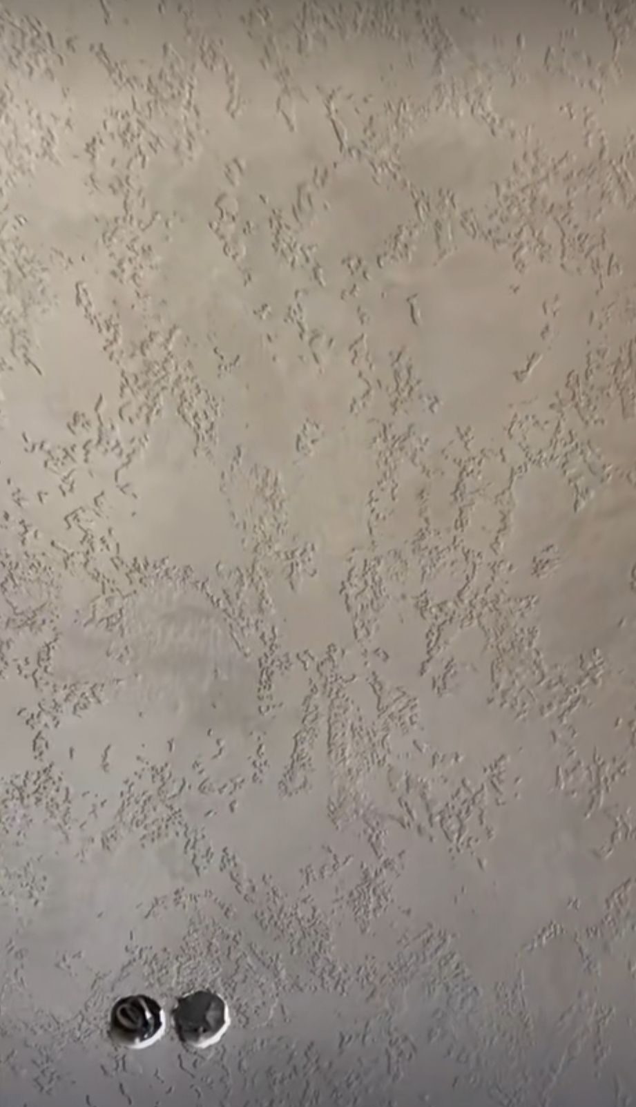 Szara ściana z fakturą dekoracyjną i widocznymi otworami na gniazdka elektryczne w dolnej części.