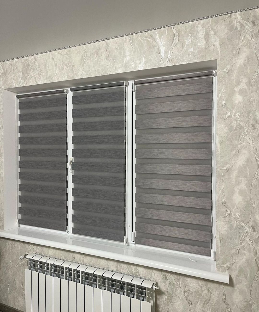 Trzyczęściowe okno z szarymi roletami dzień-noc, marmurkowy wzór na ścianie i biały grzejnik pod parapetem.