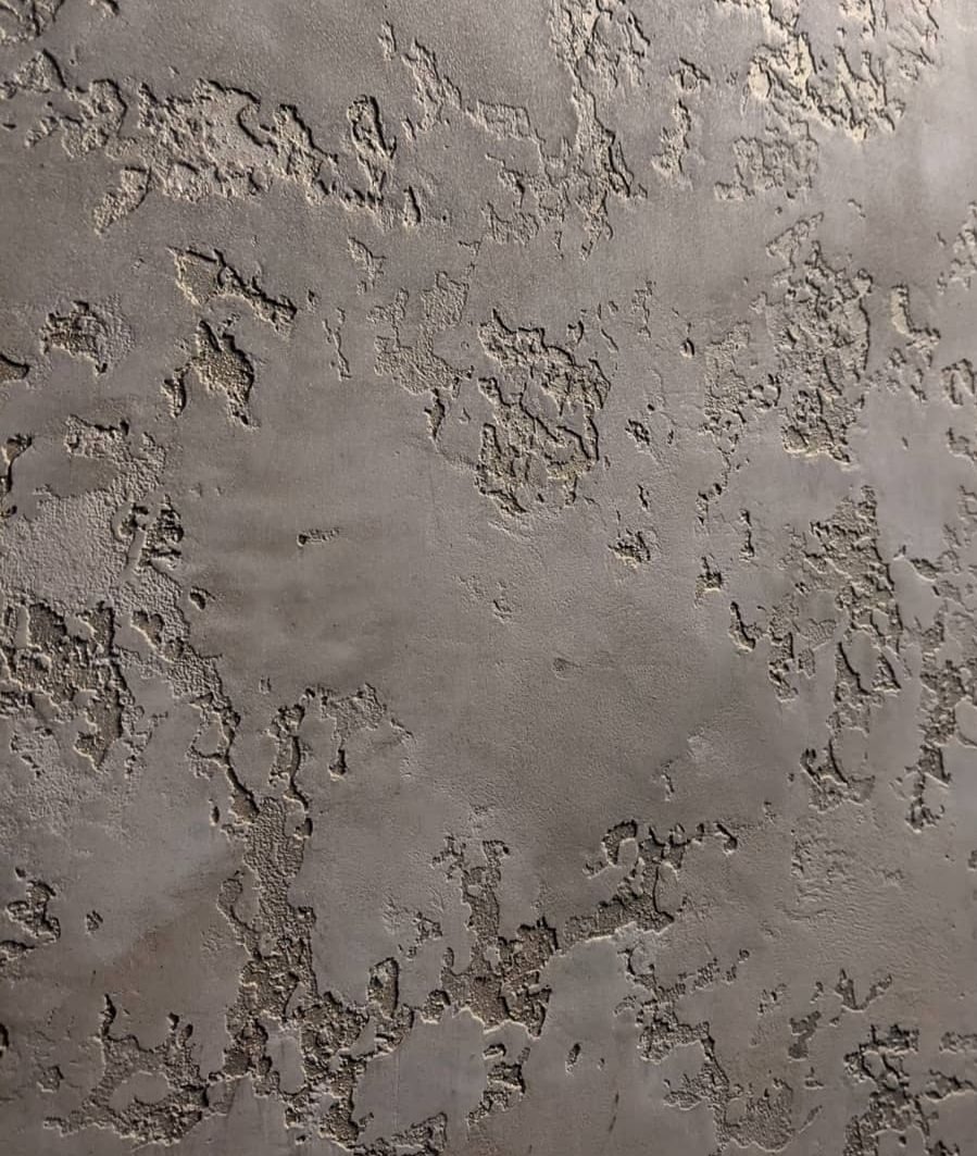 Szara ściana z dekoracyjną, nierówną fakturą imitującą spękania i ubytki, z delikatnym połyskiem w zagłębieniach.