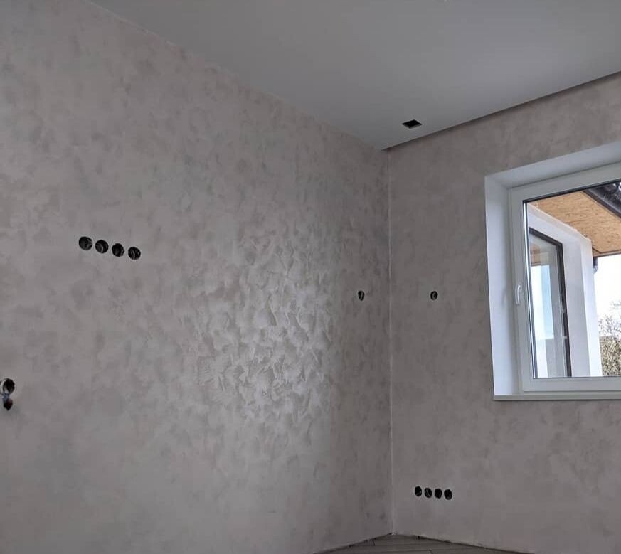 Ściana w pokoju po nałożeniu dekoracyjnej gładzi w odcieniach szarości i beżu, widoczne puste otwory na gniazdka elektryczne i białe okno.