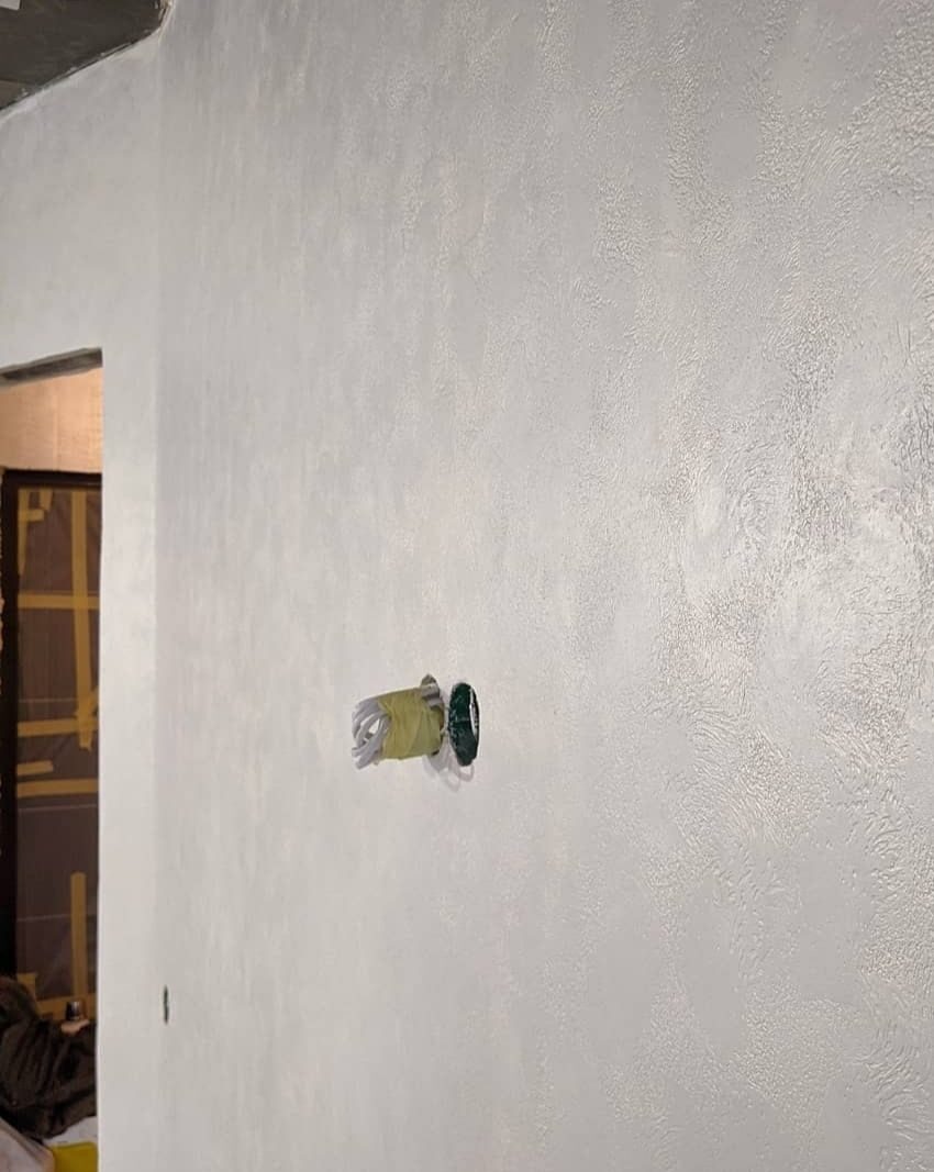 Ściana pokryta dekoracyjną, jasną gładzią, z wystającymi kablami zabezpieczonymi taśmą malarską w miejscu gniazdka elektrycznego.