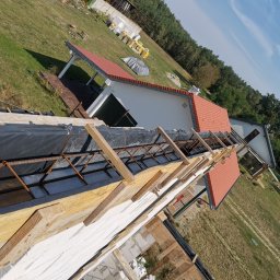 Krzyś Bud Usługi Ogólnobudowlane Krzysztof Dura - Wznoszenie muru z białych bloczków z widocznym zbrojeniem i drewnianym szalunkiem, w tle budynek z czerwoną dachówką i zielone otoczenie.