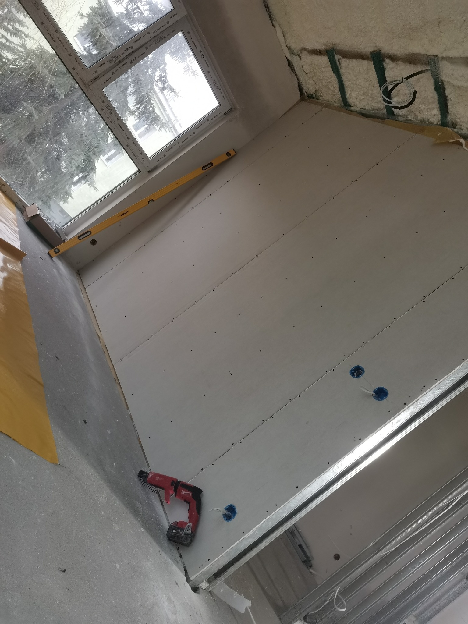 Montaż płyt kartonowo-gipsowych na ścianie, widoczny poziomica przy oknie, wkrętarka akumulatorowa na podłodze i instalacja elektryczna.