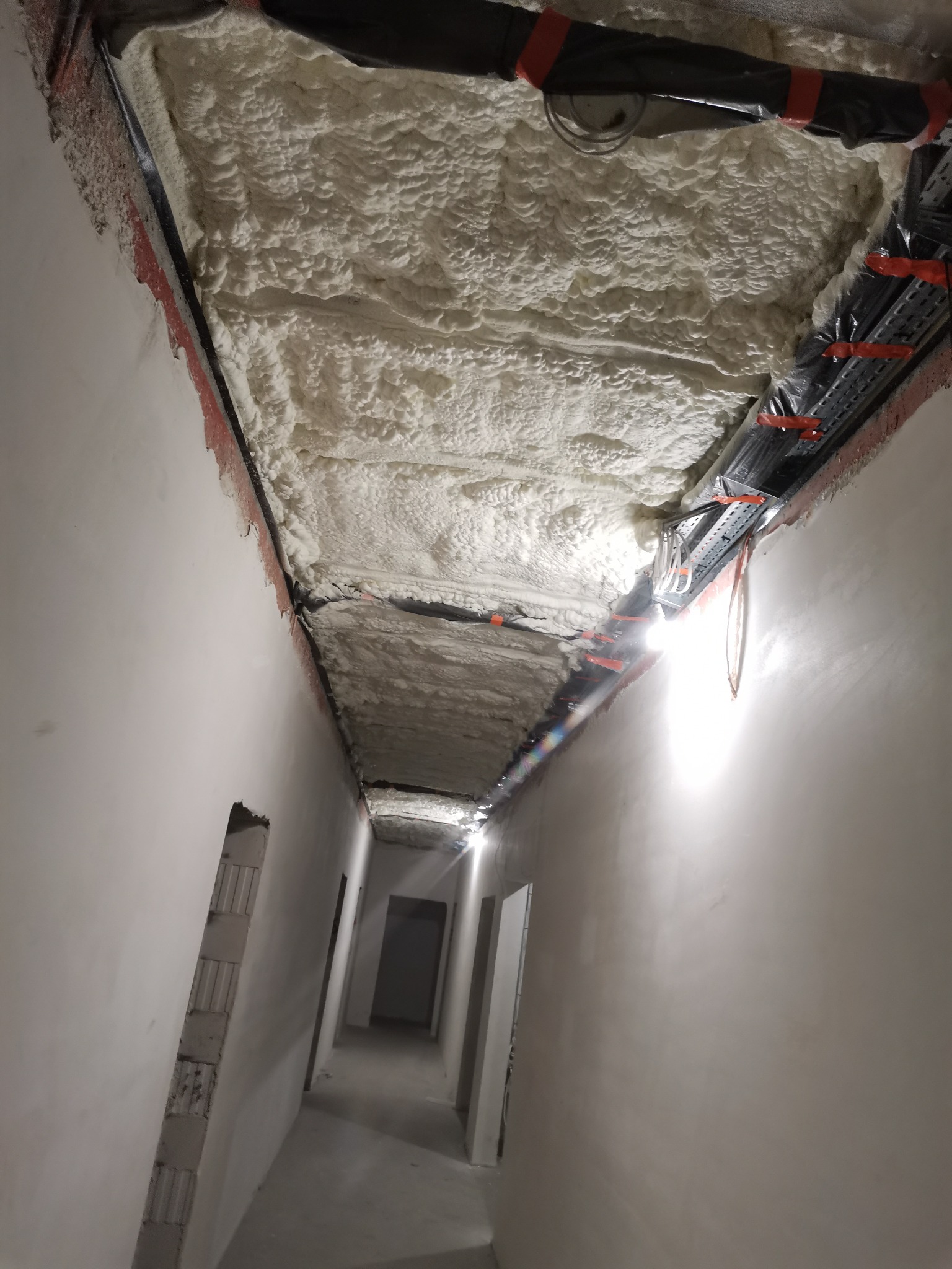 Korytarz w trakcie prac wykończeniowych z widoczną izolacją piankową na suficie, instalacjami zabezpieczonymi folią i taśmą, oraz surowymi ścianami przygotowanymi do dalszych prac.