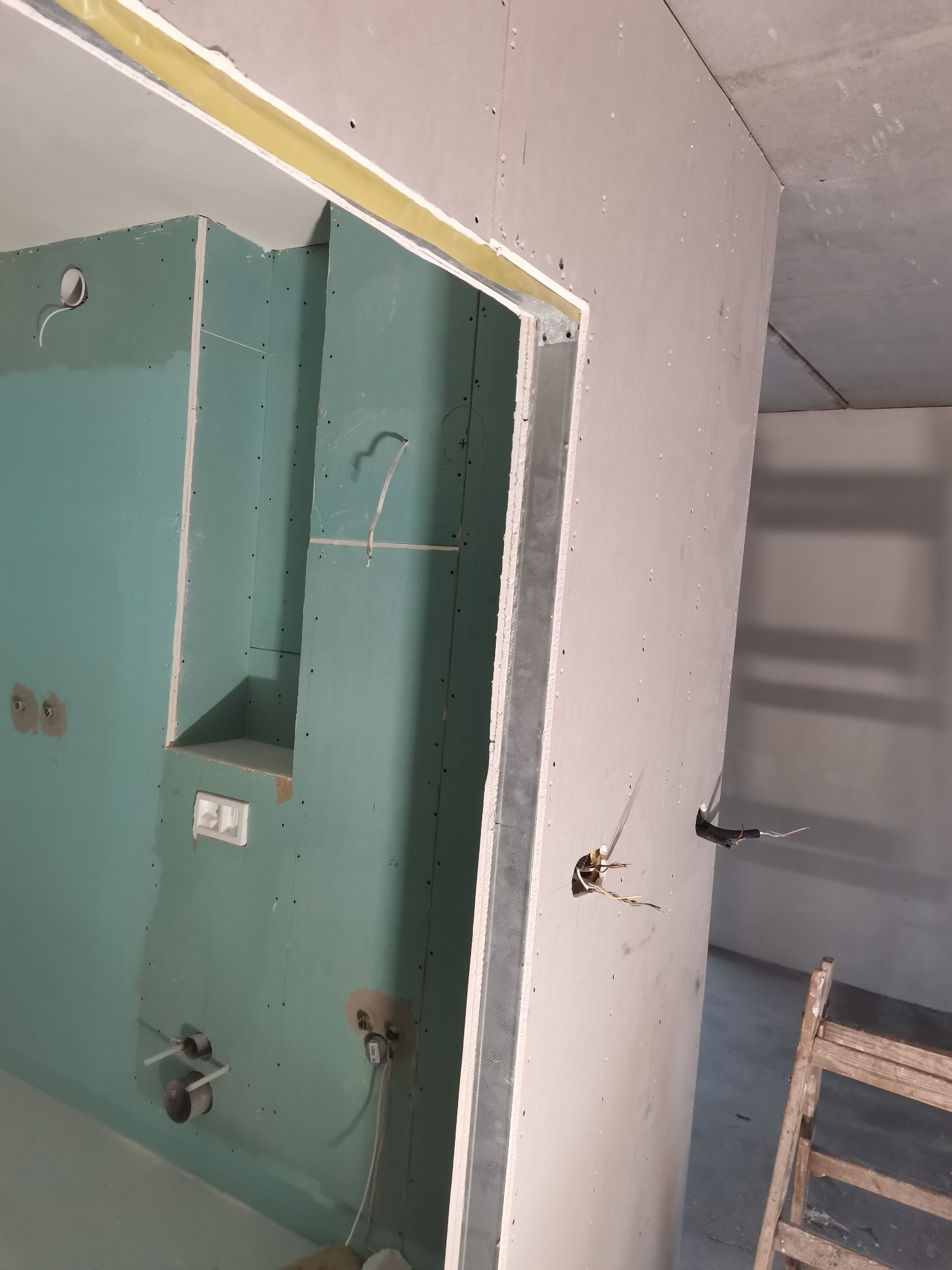 Montaż zielonej płyty gipsowo-kartonowej w łazience, widoczne otwory na instalacje wodne i elektryczne, oraz wnęka w ścianie. Zabezpieczone taśmą łączenie płyty na suficie. Fragment drabiny w rogu.