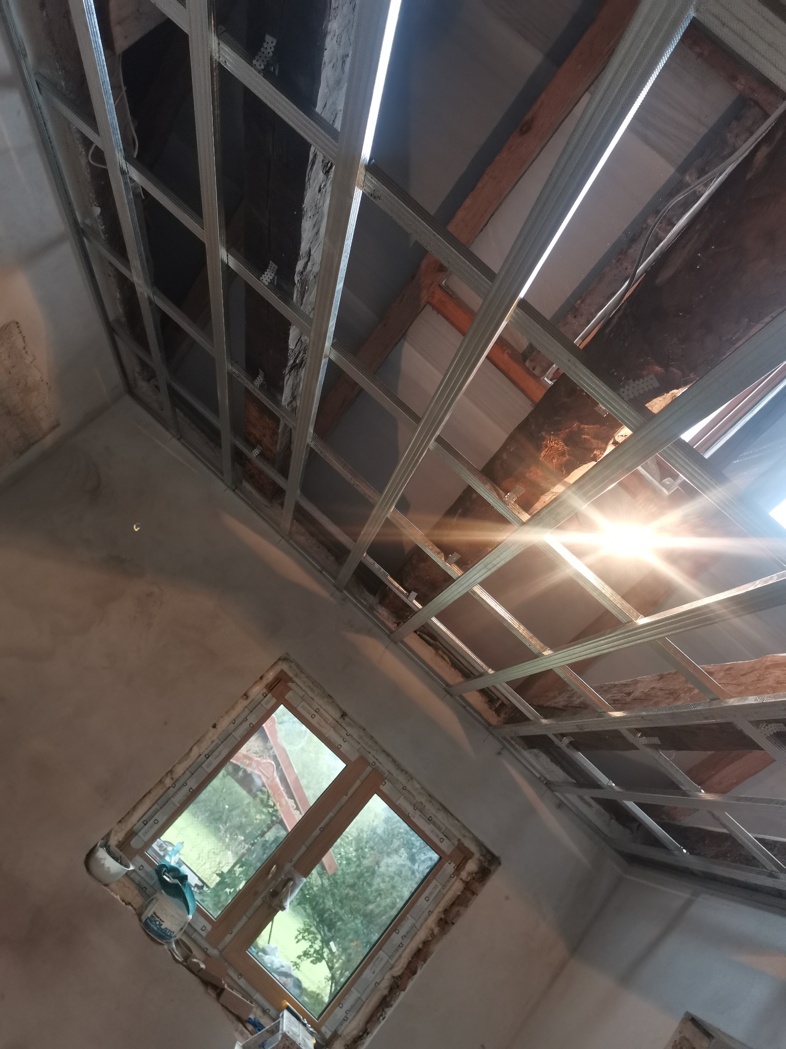 Montaż stelaża sufitu podwieszanego z profili metalowych w pomieszczeniu z oknem, widoczne elementy konstrukcji dachu i niedokończone ściany.