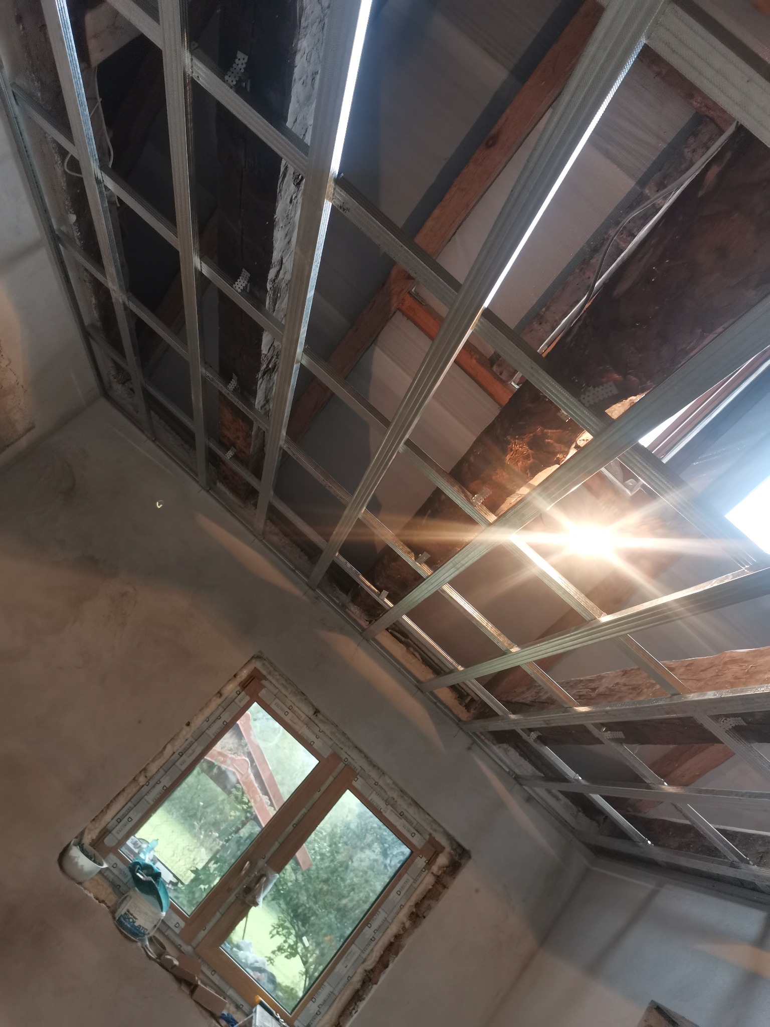 Metalowa konstrukcja sufitu podwieszanego z widocznymi elementami drewnianymi, oświetlona wpadającym przez otwór w stropie światłem, z oknem w ścianie z widokiem na zieleń.