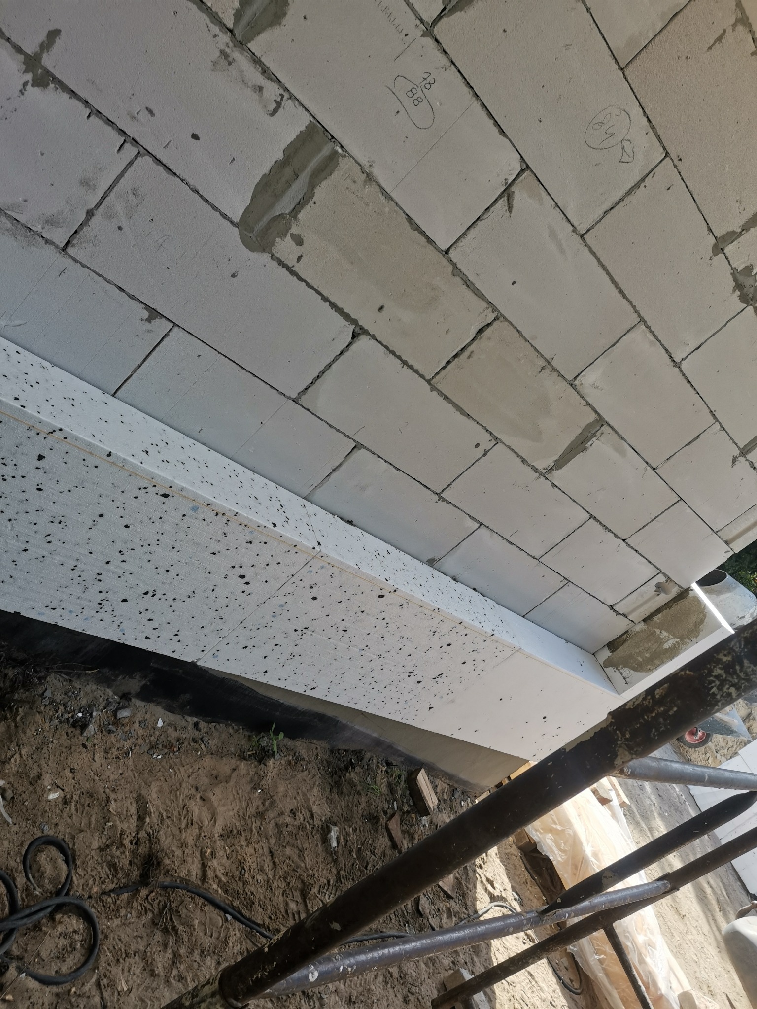 Ściana z białych bloczków z widocznymi śladami zaprawy i odręcznymi notatkami na niektórych elementach, ocieplona styropianem, widok z dołu z rusztowaniem w Brączewie.