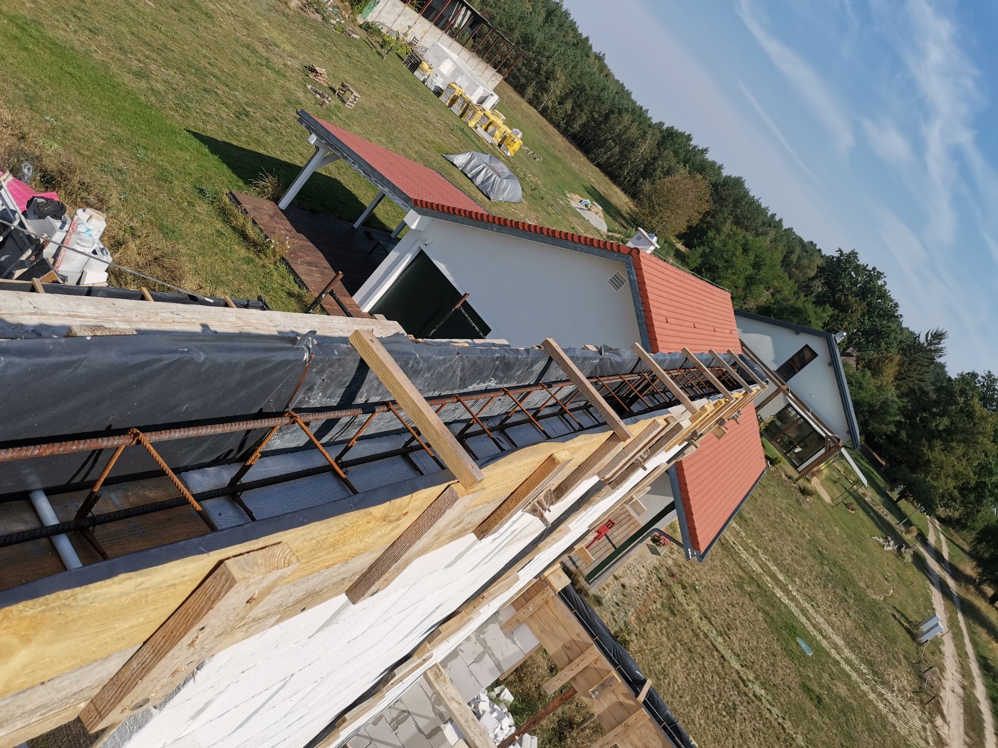 Wznoszenie muru z białych bloczków z widocznym zbrojeniem i drewnianym szalunkiem, w tle budynek z czerwoną dachówką i zielone otoczenie.
